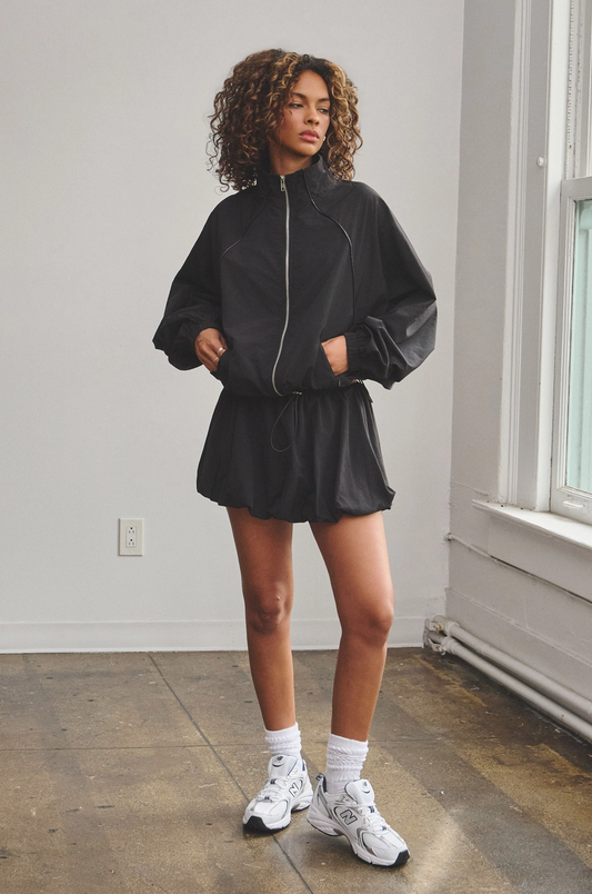 A female model wearing a black zip-up track jacket and a matching bubble mini skort with an adjustable drawstring.