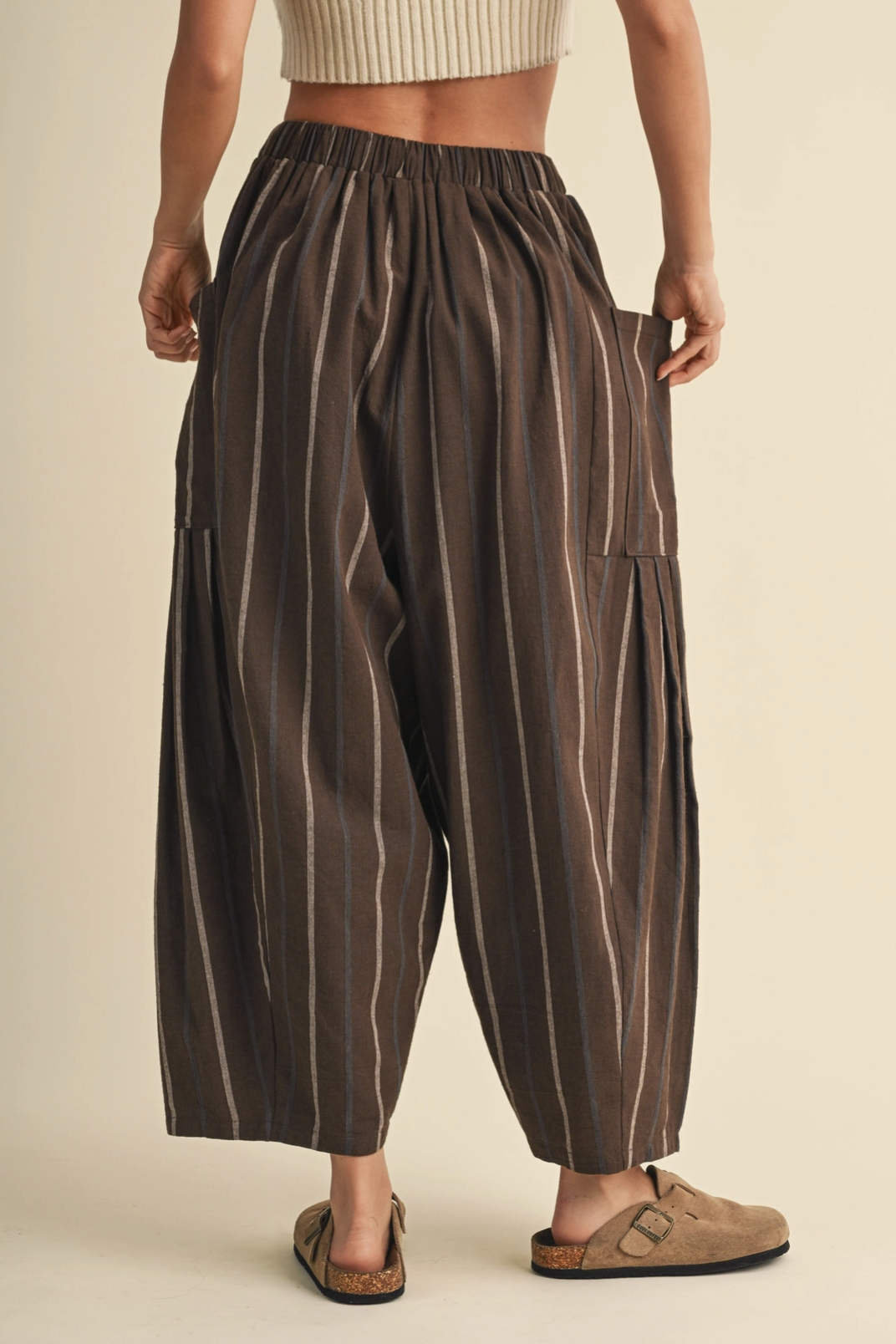 A person wearing loose, dark brown striped pants with side pockets, paired with tan slide sandals and a ribbed beige crop top, shown from the waist down against a plain cream background.