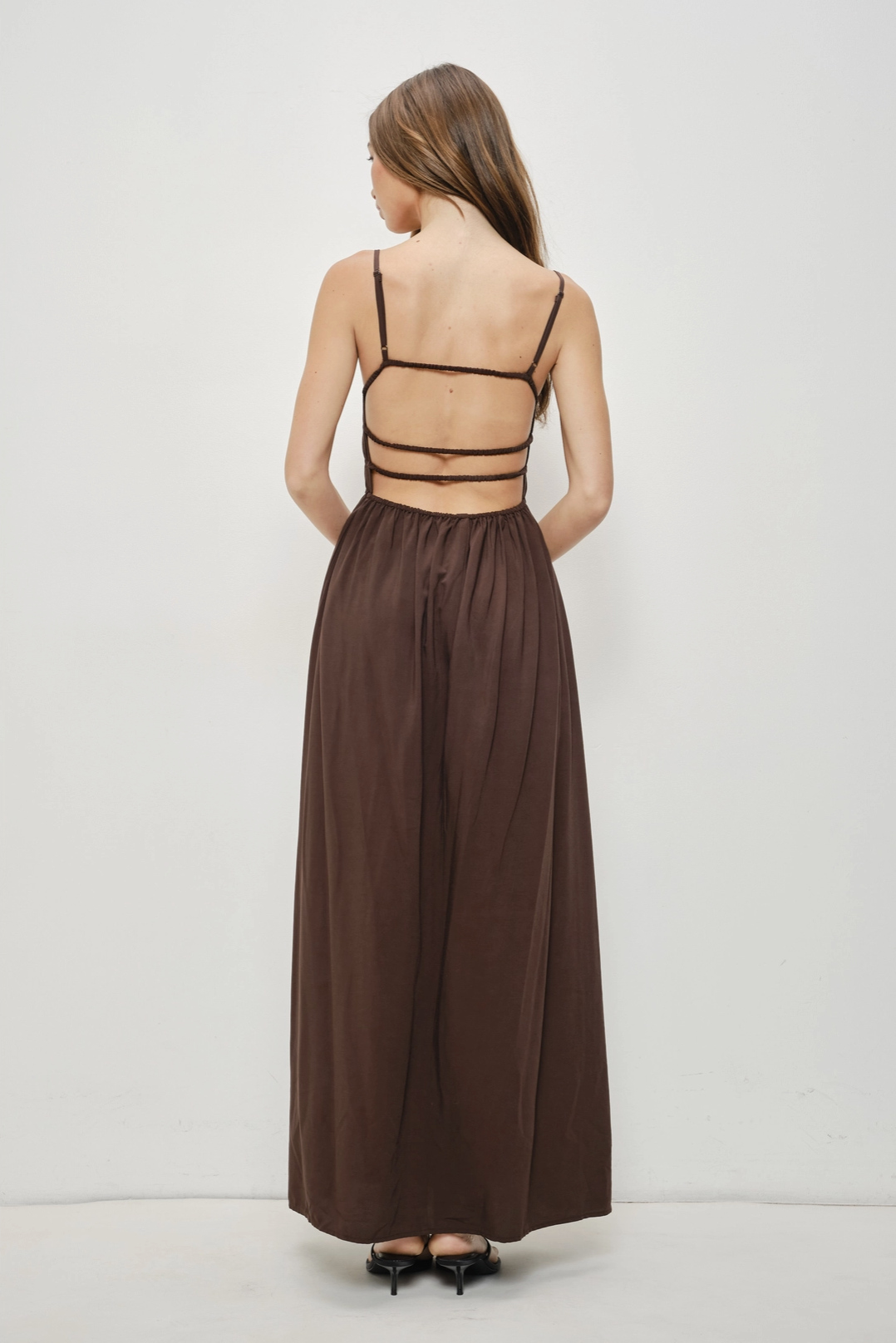 A woman viewed from the back wearing a brown maxi dress with thin black spaghetti straps, a strappy open back design, and a high-waisted flowy skirt, paired with black heels against a plain white studio background.