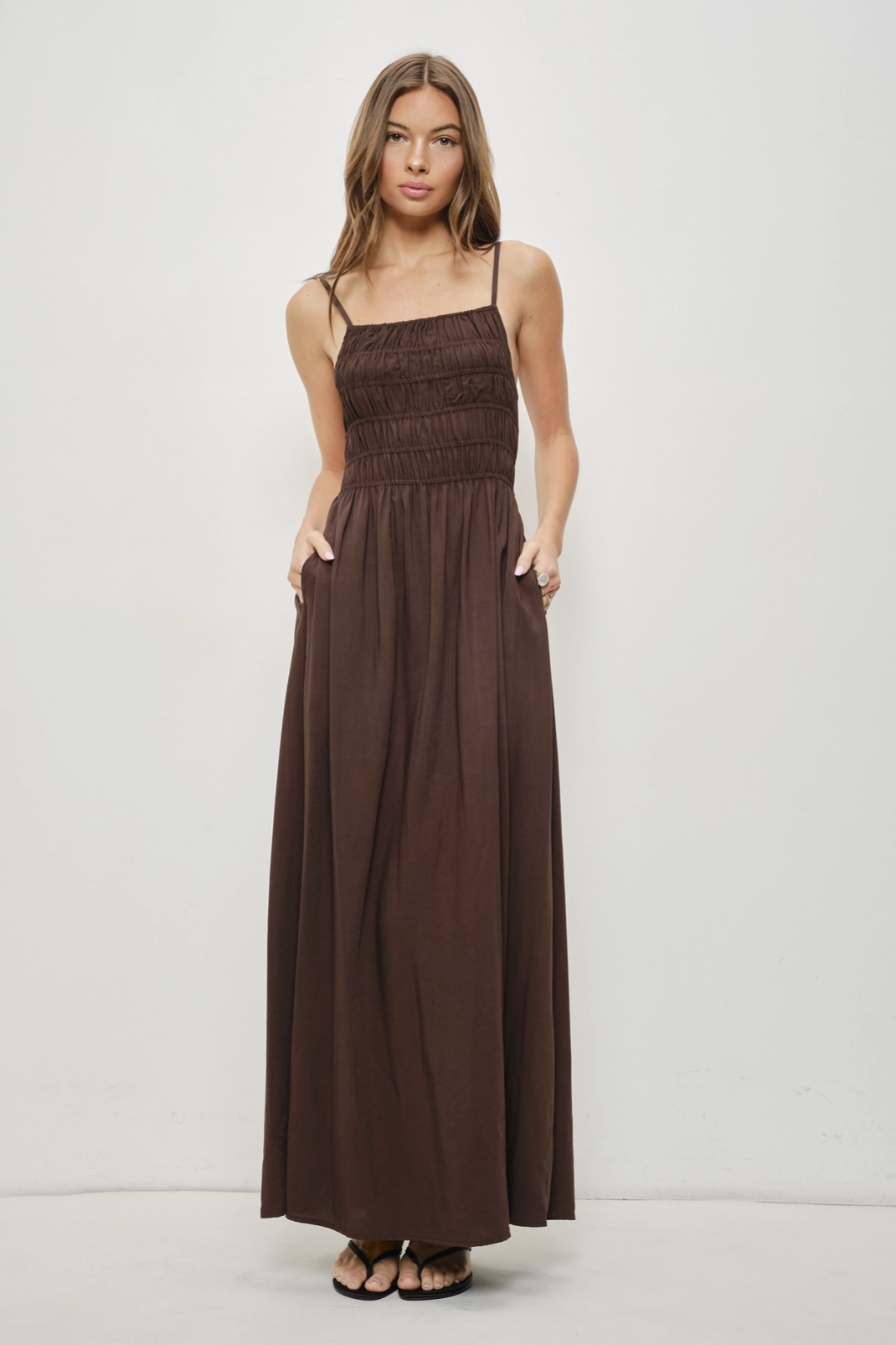 A woman modeling a dark brown maxi dress with thin spaghetti straps, a smocked bodice, and side pockets, standing against a plain white studio background.