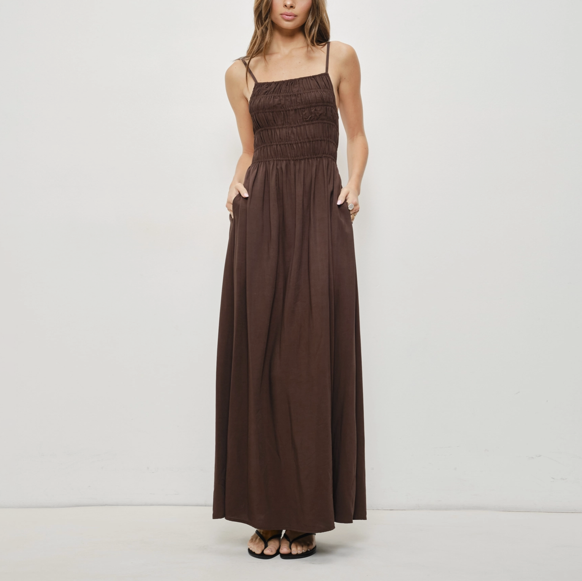 A woman modeling a dark brown maxi dress with thin spaghetti straps, a smocked bodice, and side pockets, standing against a plain off-white studio background.