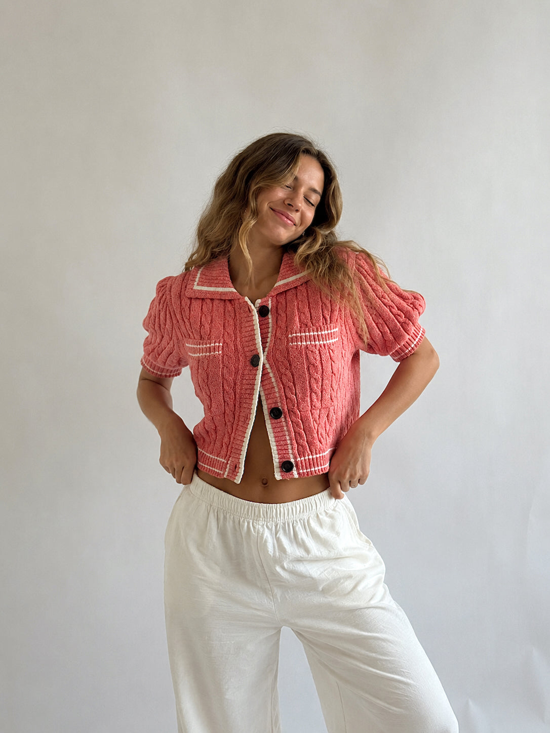 A woman modeling a short-sleeve pink cable-knit cropped jacket with white contrast trim and white buttons, paired with white pants, against a plain light gray studio background.