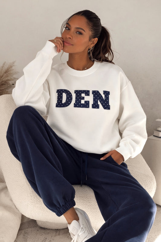 Woman wearing a white sweatshirt with 'DEN' printed on it, sitting on a chair.