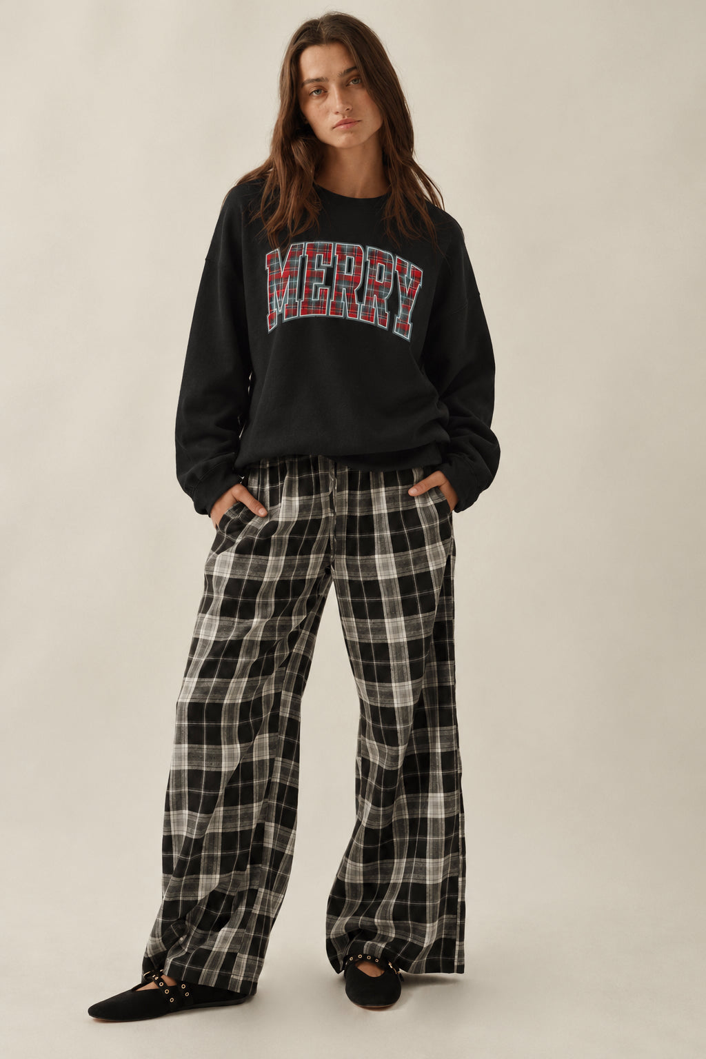 A woman modeling a black crewneck sweatshirt with a red and blue plaid 'MERRY' graphic across the chest, paired with black and white plaid wide-leg pants, against a plain beige studio background.