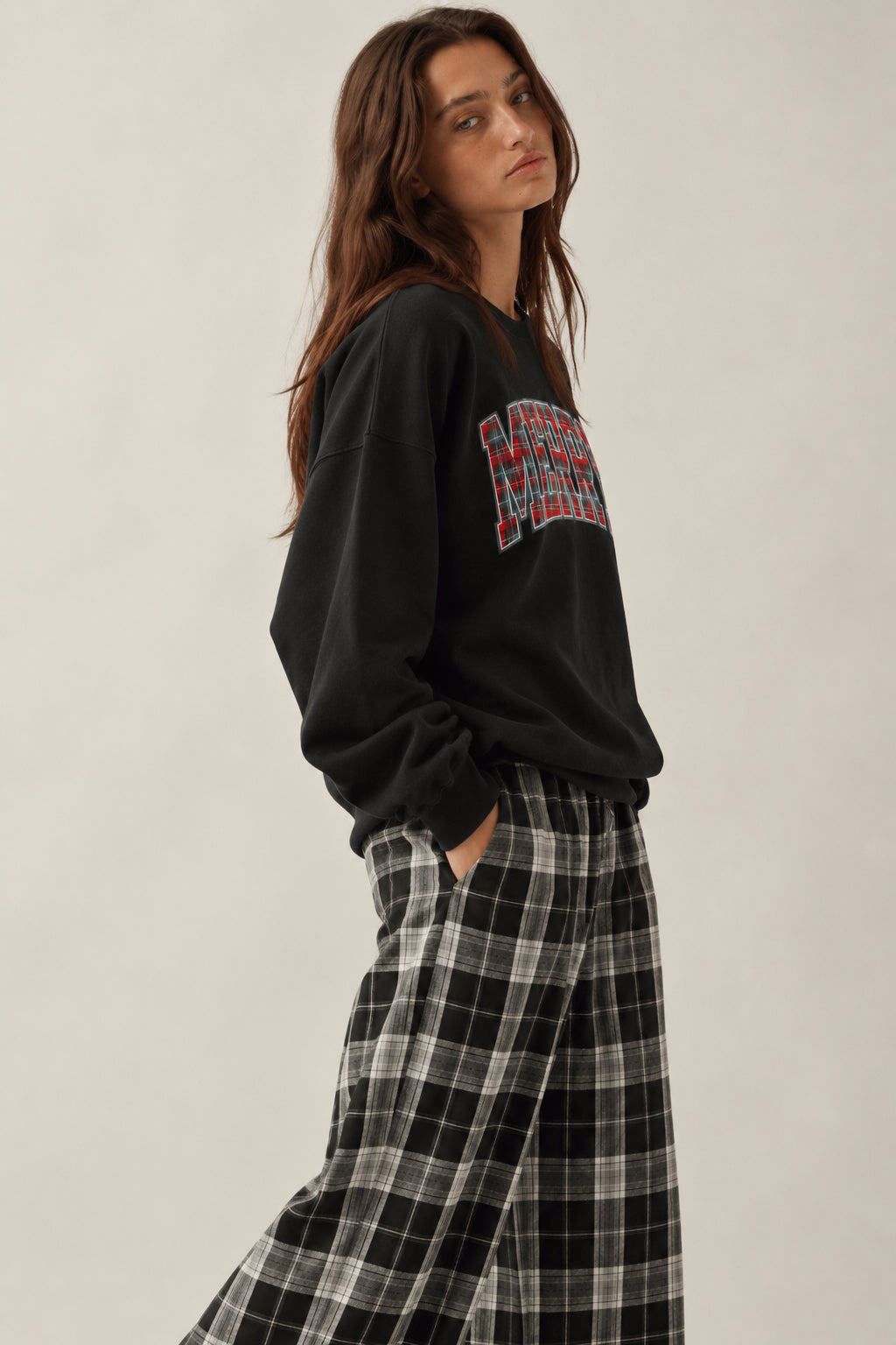 A woman wearing a black crewneck sweatshirt with a red and blue graphic on the chest, paired with black and white plaid wide-leg pants, photographed against a plain light beige studio background.