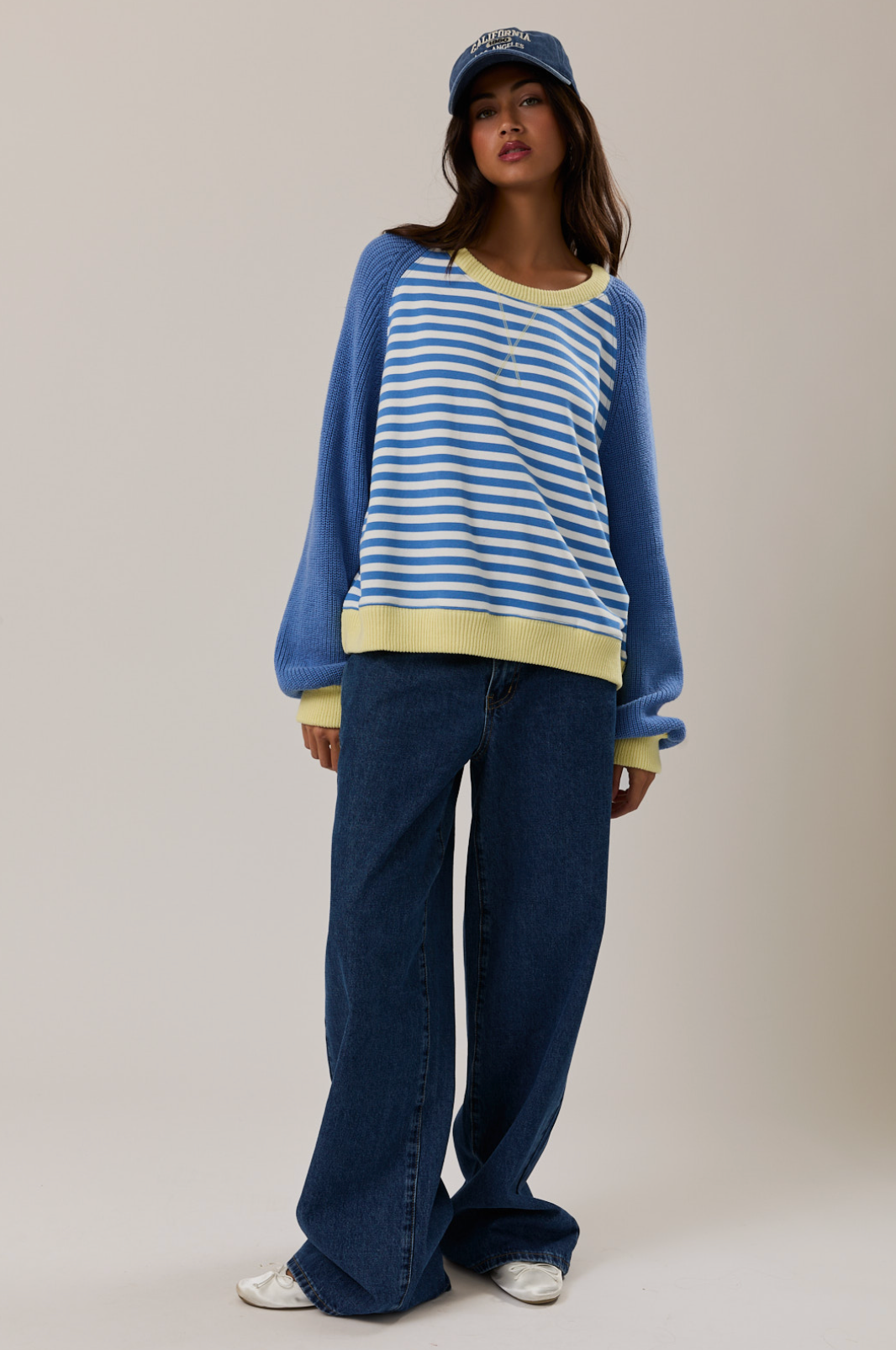 Woman wearing a blue and white striped sweater with a yellow shirt underneath, paired with blue jeans and a blue cap on a plain background.