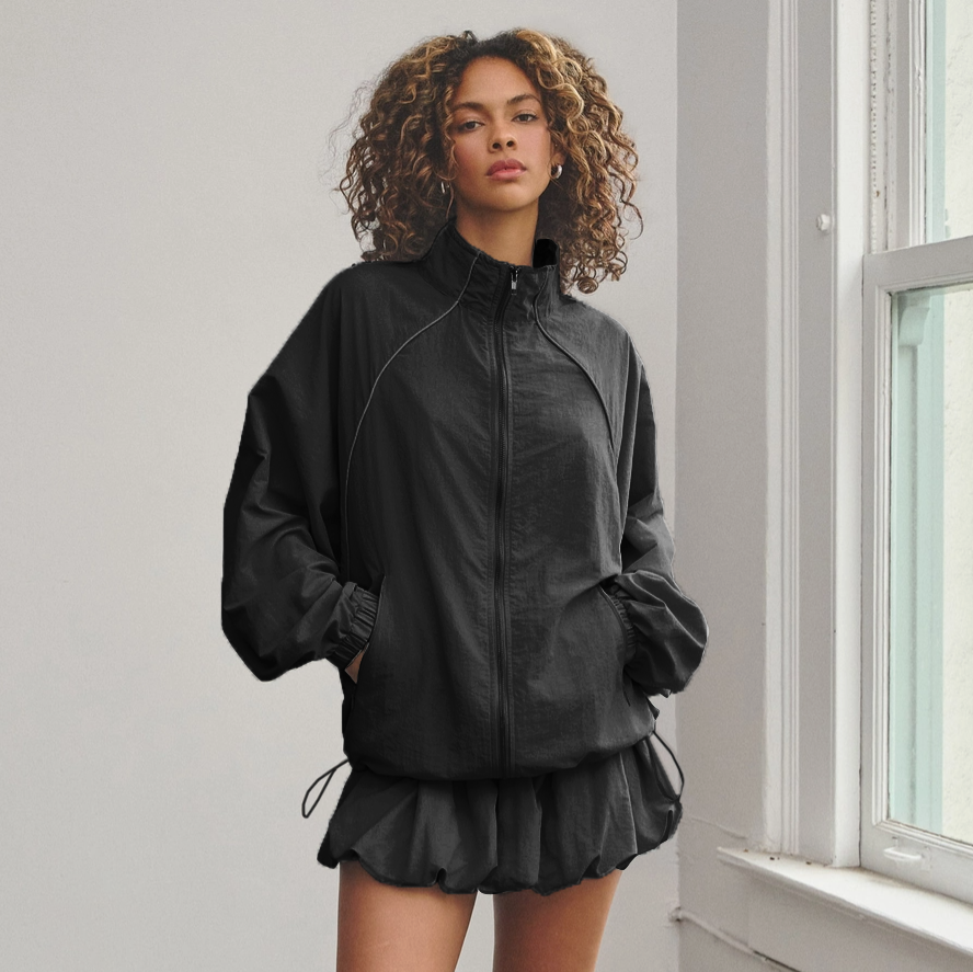 A female model wearing a black zip-up track jacket and a matching bubble mini skort with an adjustable drawstring.