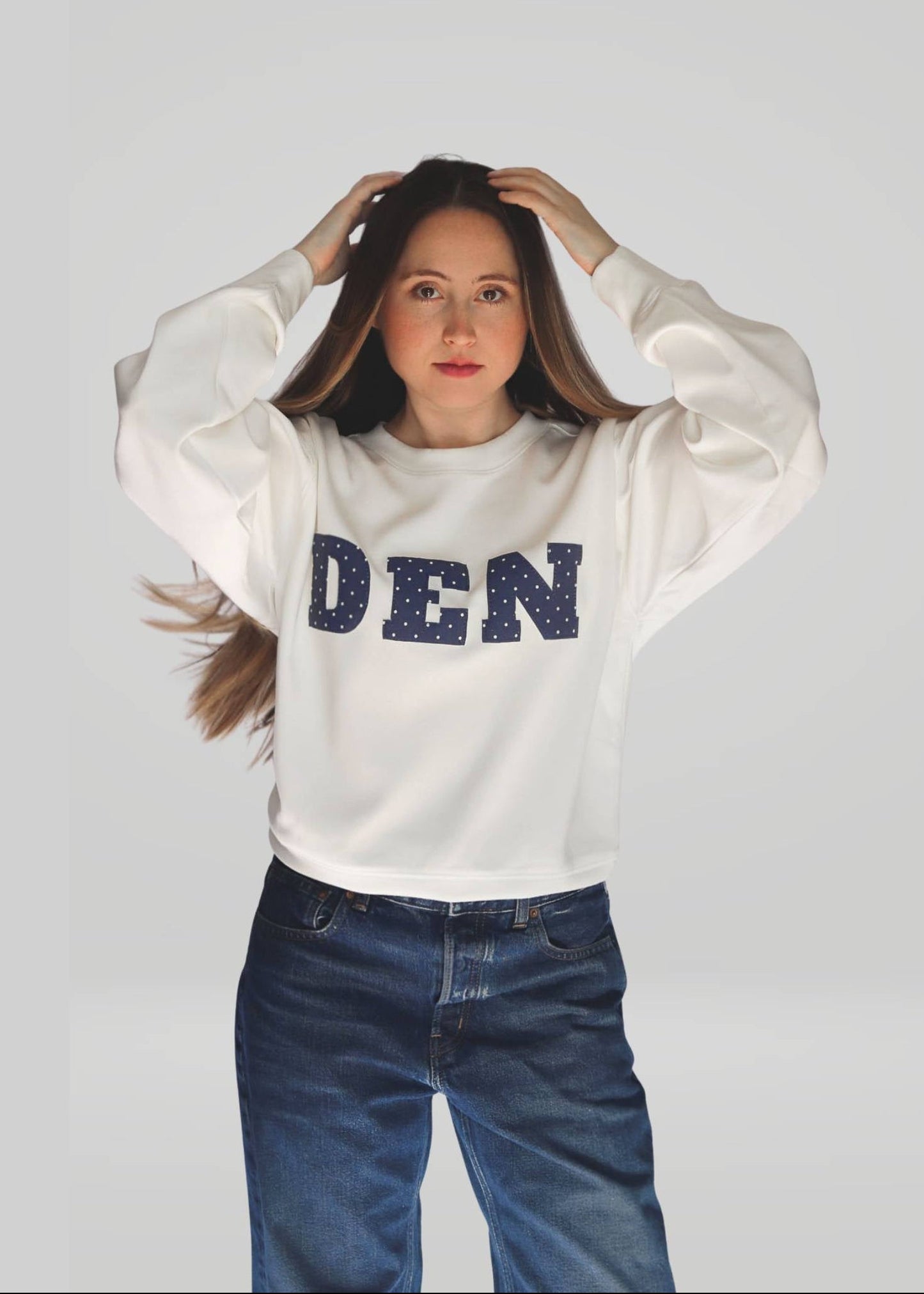 Person wearing a white sweatshirt with 'DEN' printed on it against a plain background