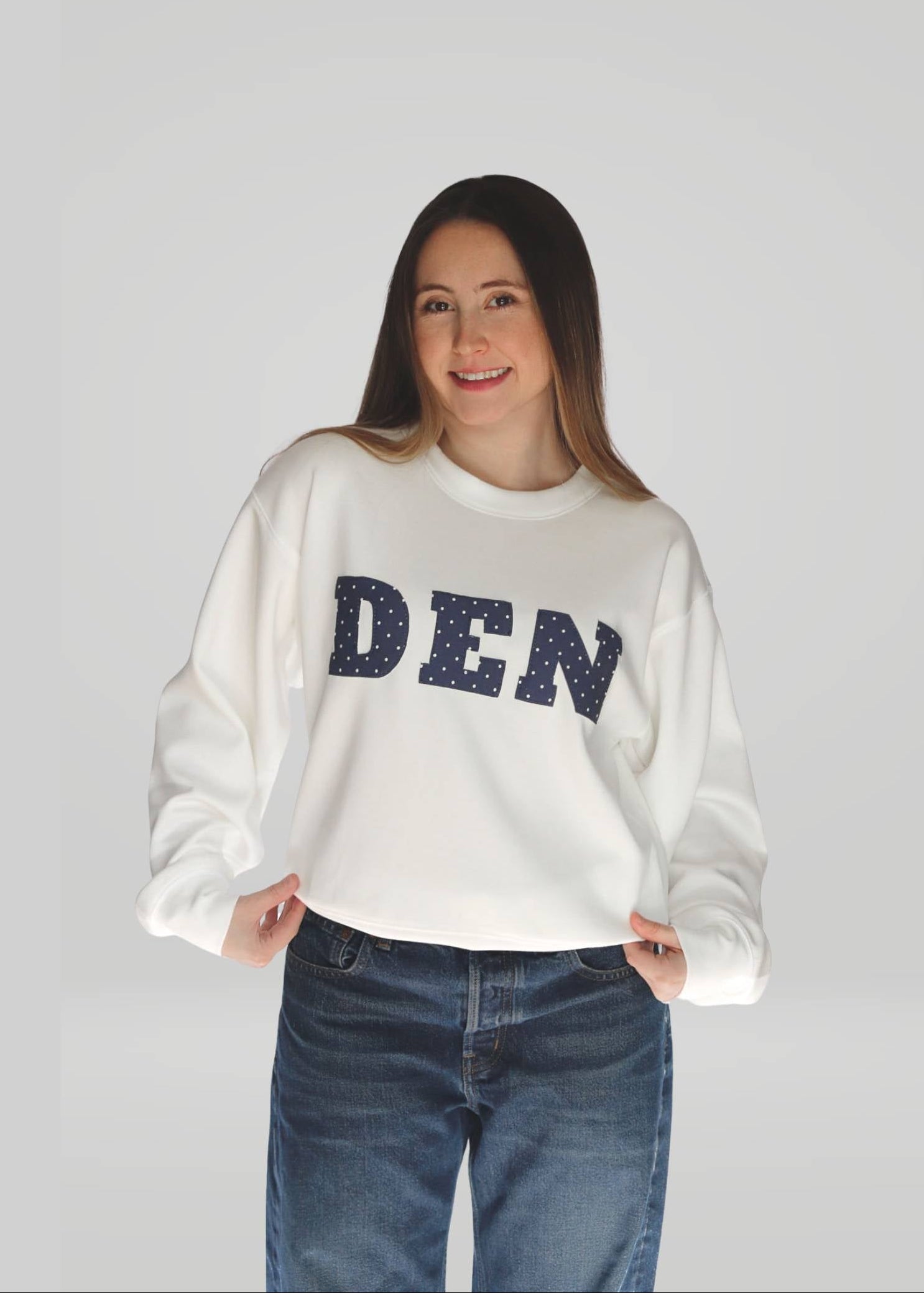 Woman wearing a white sweatshirt with 'DEN' on it against a plain background