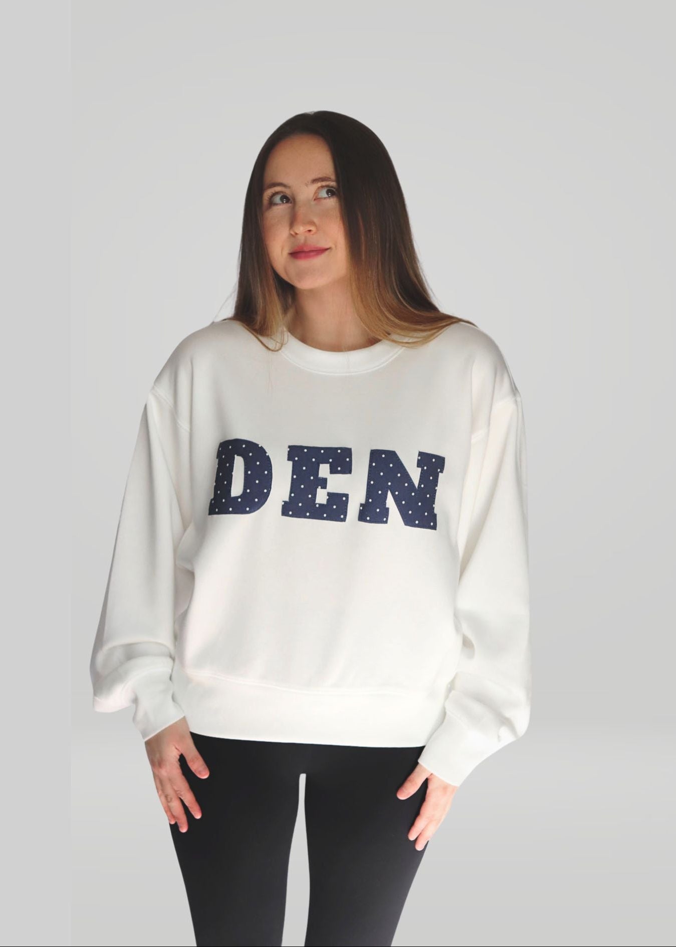 Woman wearing a white sweatshirt with 'DEN' printed on it against a plain background