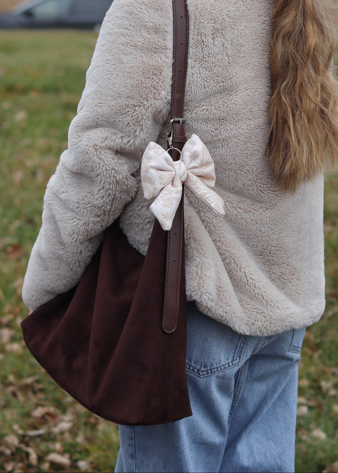 Person wearing a beige fleece jacket with a brown bag featuring a white bow, standing outdoors.