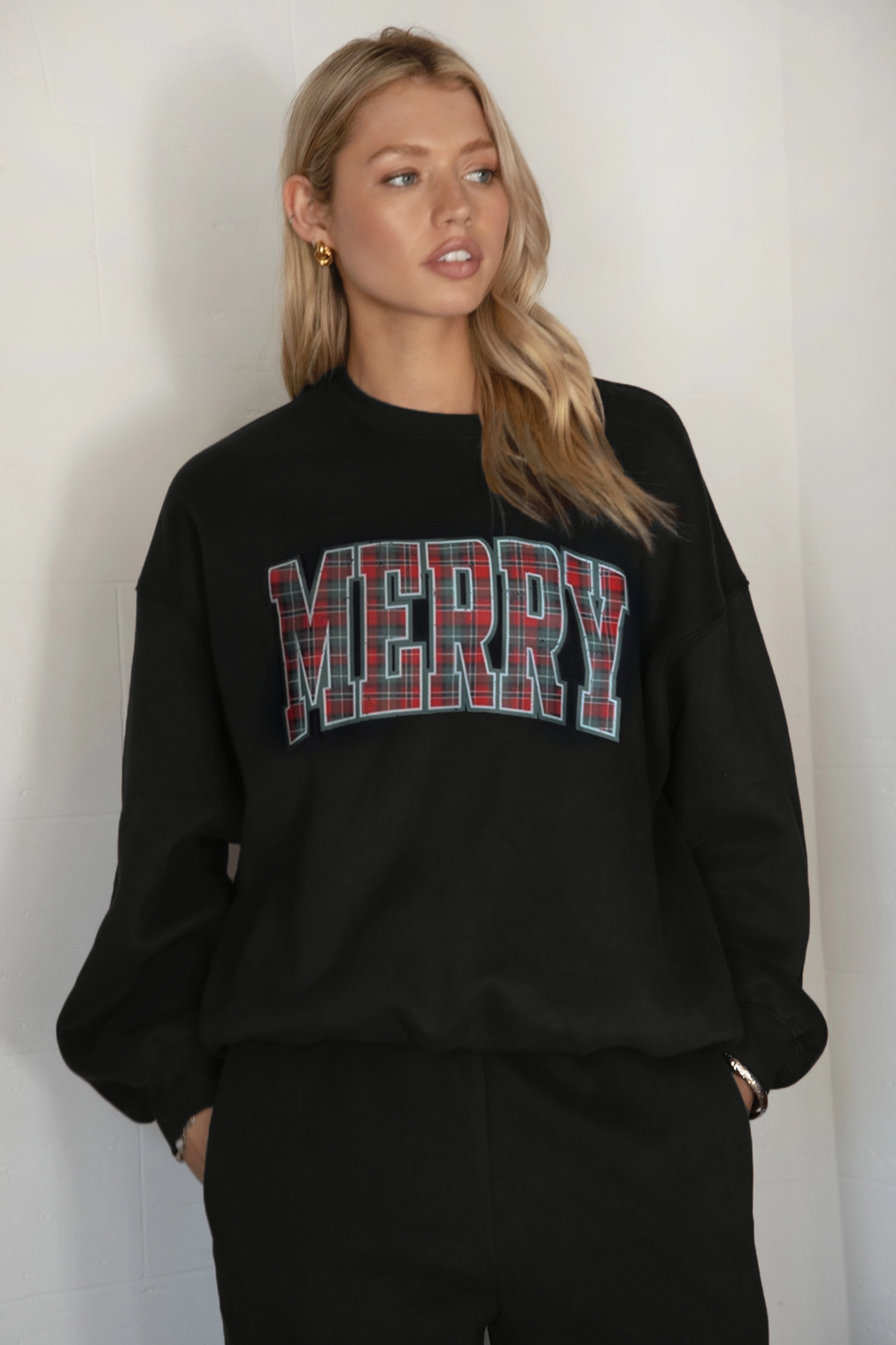 Person wearing a black sweatshirt with 'MERRY' in plaid letters against a plain background