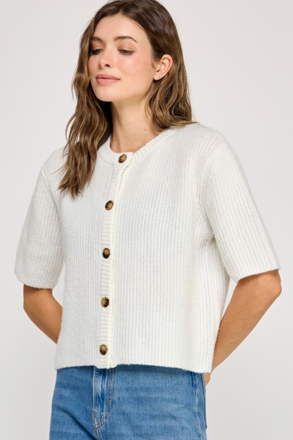 Woman wearing a white ribbed knit cardigan with brown buttons on a plain background