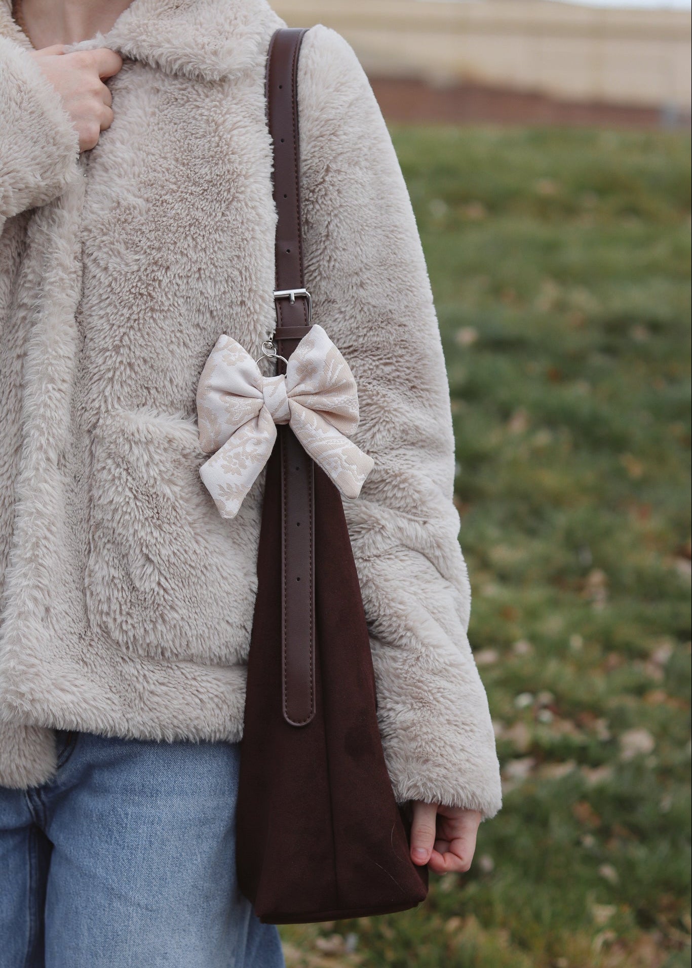 Person wearing a beige fleece jacket with a brown bag featuring a large white bow, standing outdoors.