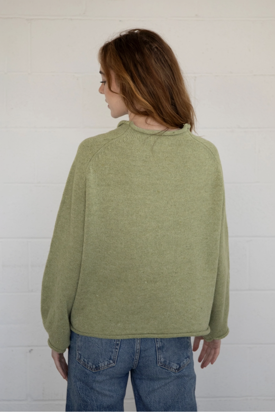 Person wearing a green sweater and blue jeans against a white wall