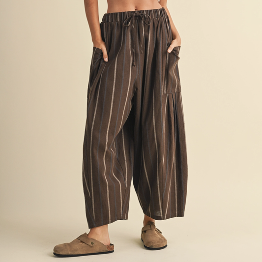 A person wearing loose brown drawstring pants with vertical beige stripes and side pockets, paired with tan slip-on shoes, against a plain beige studio background.