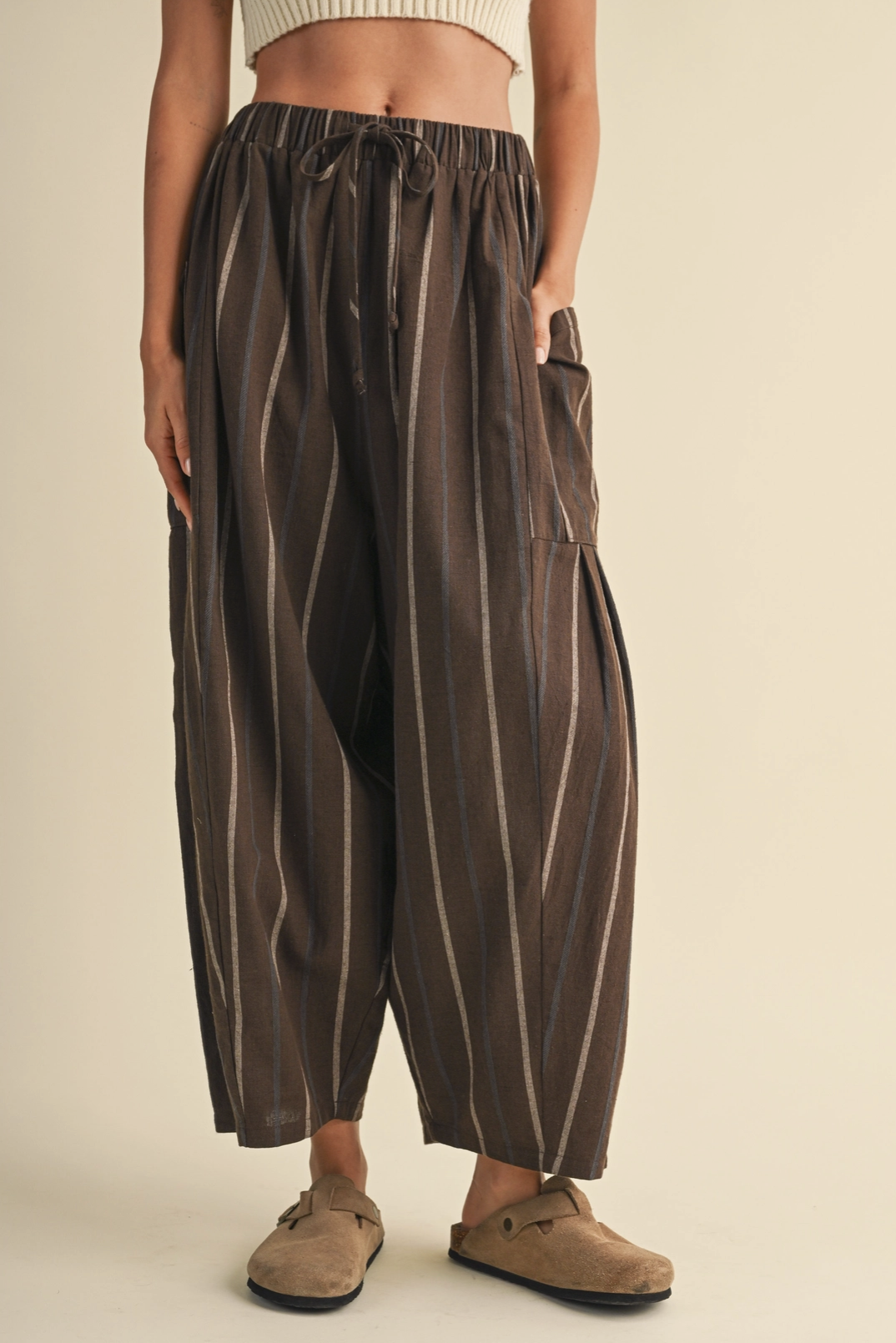 A person wearing loose, wide-leg brown pants with vertical cream stripes, an elastic drawstring waist, and side pockets, paired with tan slip-on shoes against a plain beige studio background.