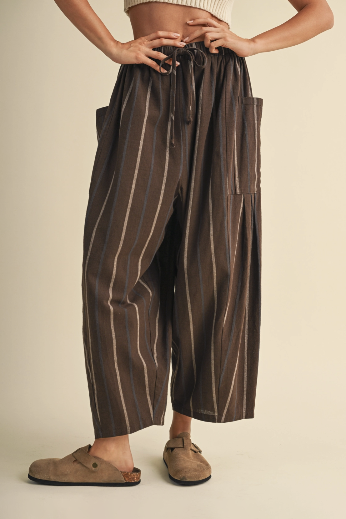 A person wearing loose, wide-leg brown pants with vertical beige stripes, a drawstring waist, and large side pockets, paired with taupe slip-on shoes against a plain cream background.