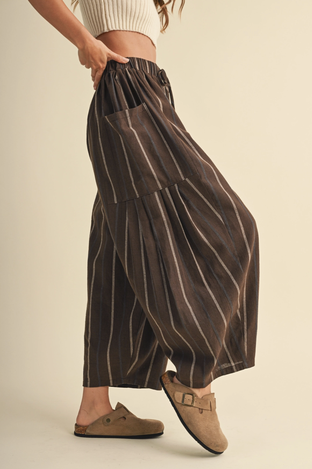 A person wearing loose, wide-leg brown pants with thin vertical cream stripes, paired with tan slip-on shoes, photographed against a plain warm beige studio background.