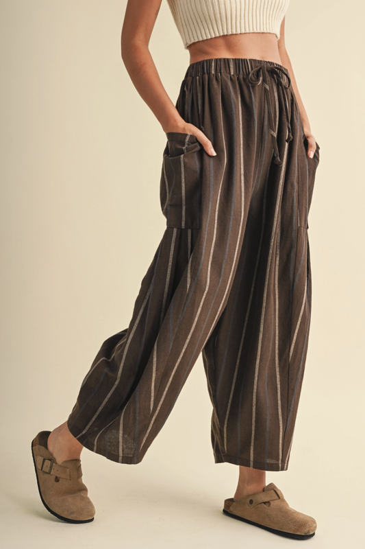 A person wearing loose, wide-leg brown pants with vertical beige stripes, side cargo pockets, and an elastic drawstring waist, paired with a cream ribbed crop top and tan clogs against a plain beige studio background.