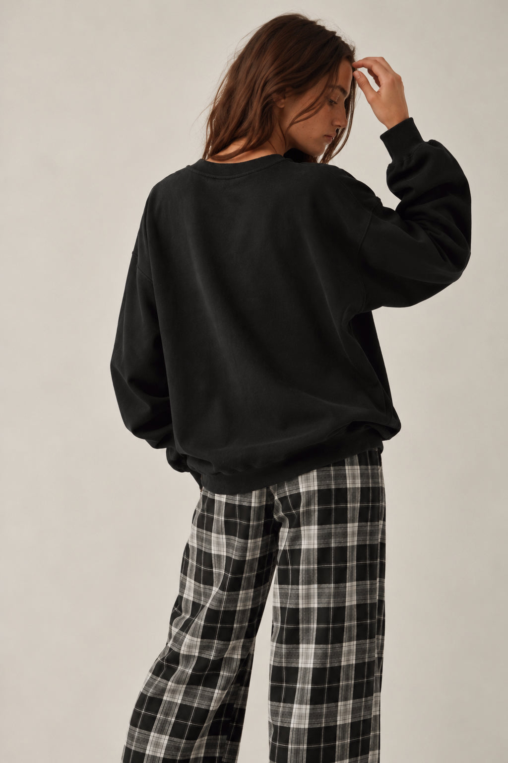 A woman wearing a black crewneck sweatshirt and black-and-white plaid pants, photographed from behind against a plain light gray studio background.