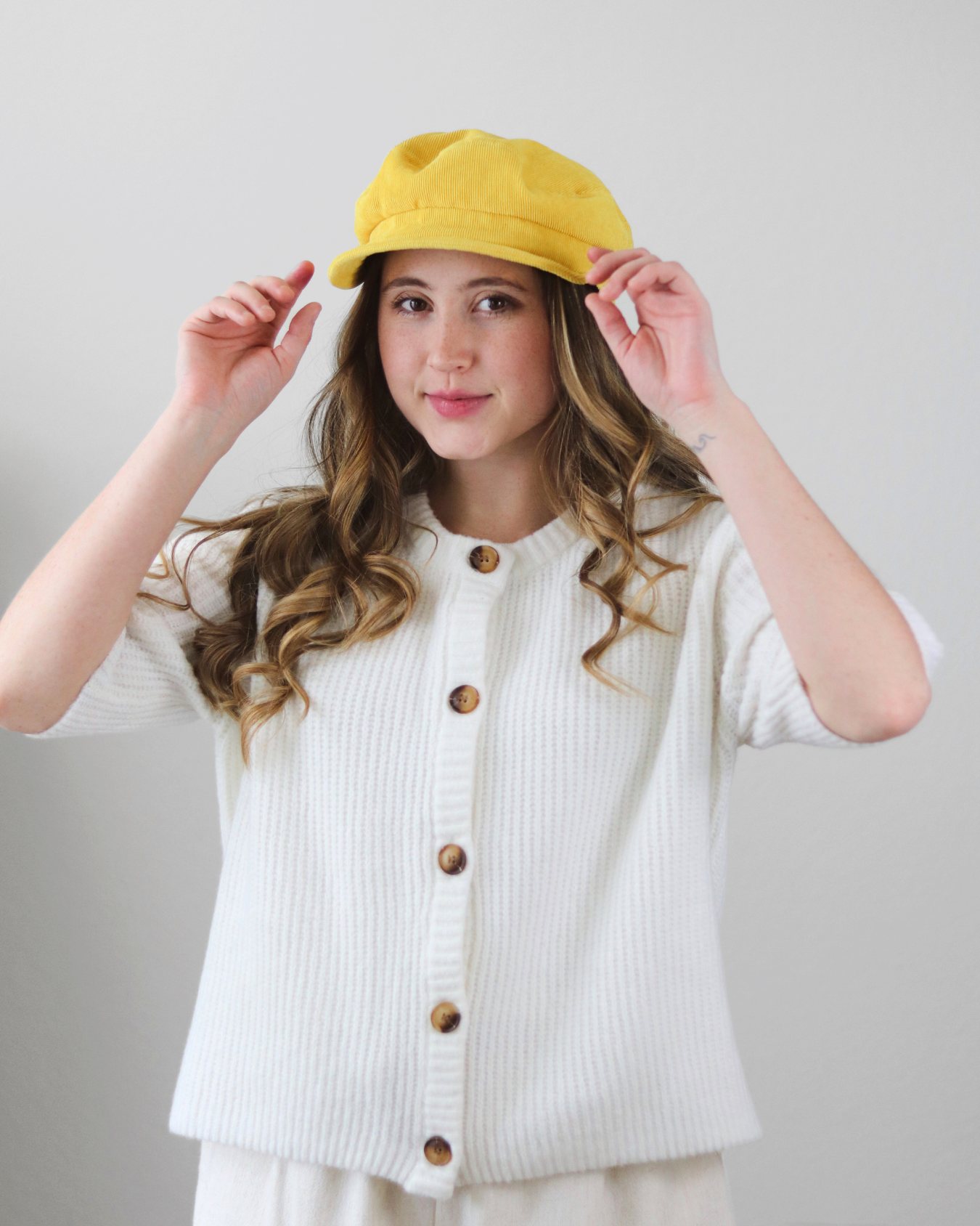 Woman wearing a yellow cap and white cardigan with brown buttons on a plain background