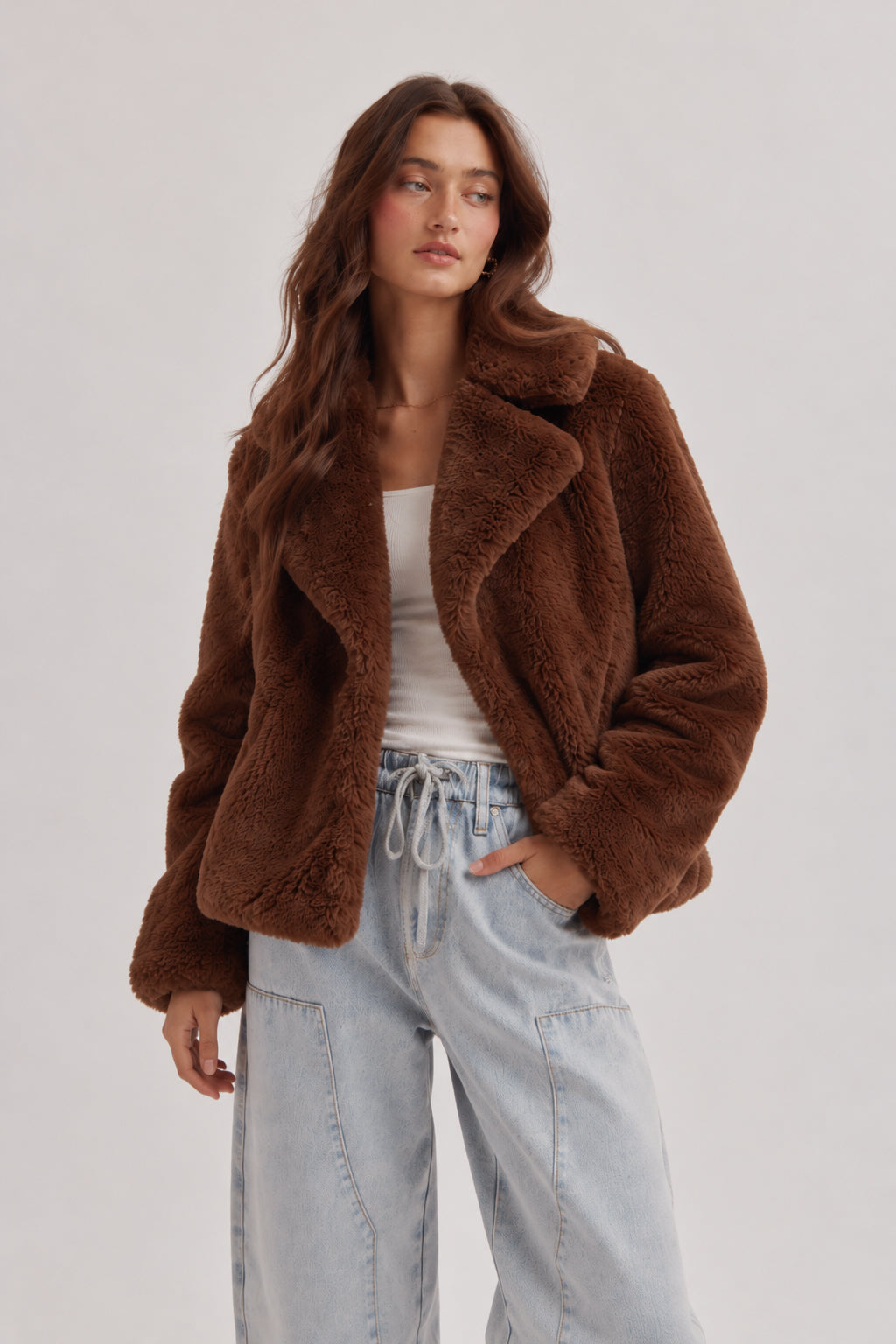 A woman modeling a brown plush teddy fleece jacket over a white tank top and light-wash wide-leg jeans, photographed against a plain light gray studio background.