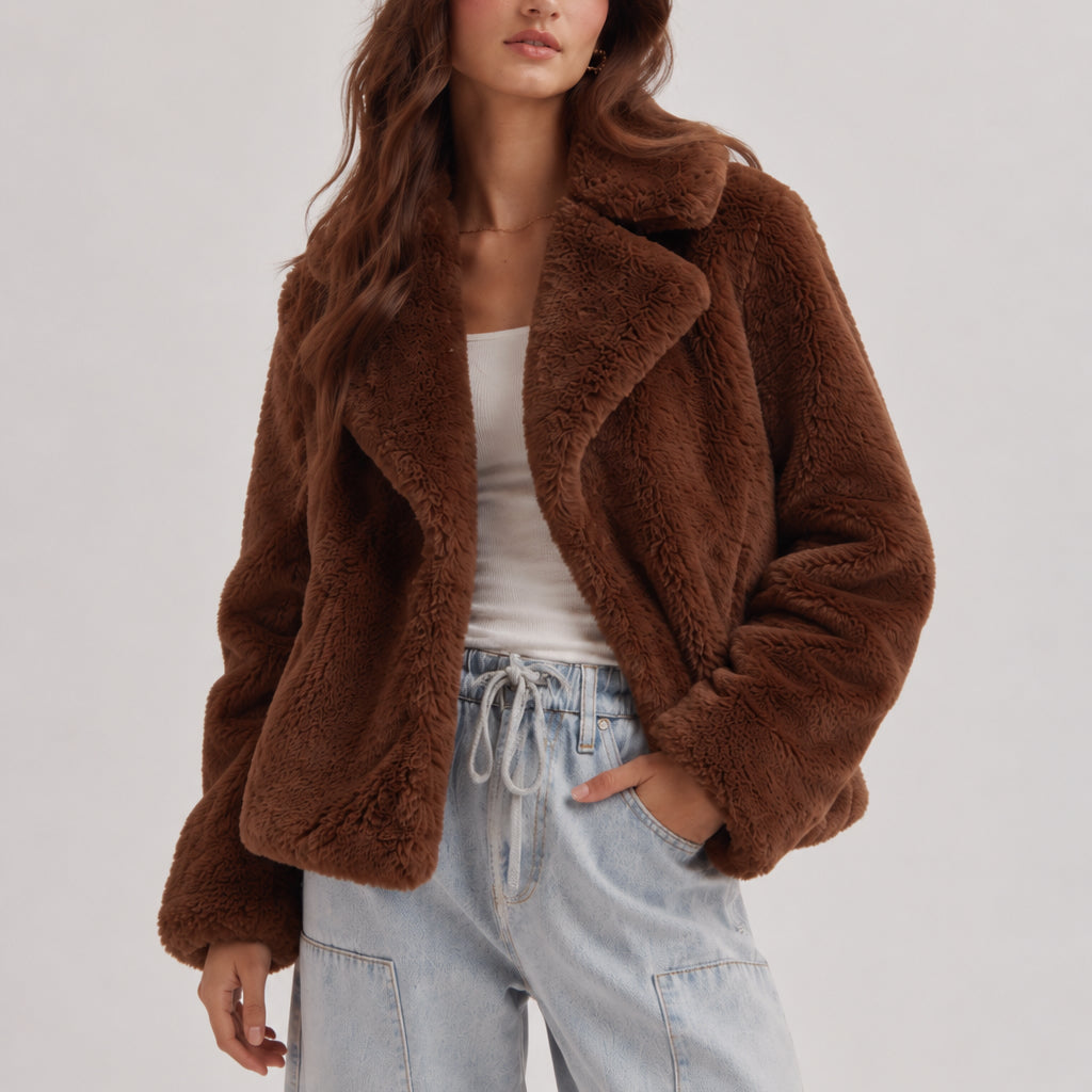 A woman wearing a brown plush teddy faux-fur jacket over a white tank top and light blue jeans, photographed against a clean light gray studio background.