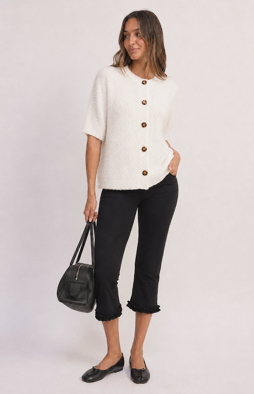 A woman modeling a short-sleeve white textured knit top with brown buttons, paired with black cropped pants and black flats, holding a black handbag against a light gray studio background.