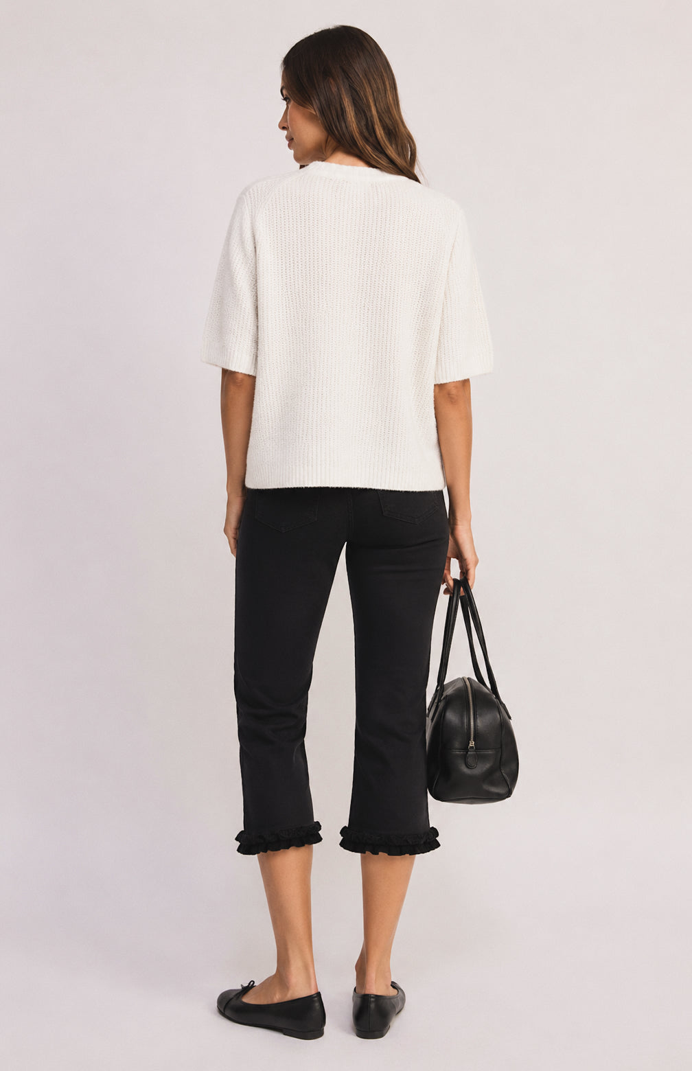 A woman shown from the back wearing a white ribbed short-sleeve top and black cropped pants with ruffled hems, holding a black handbag against a light gray studio background.