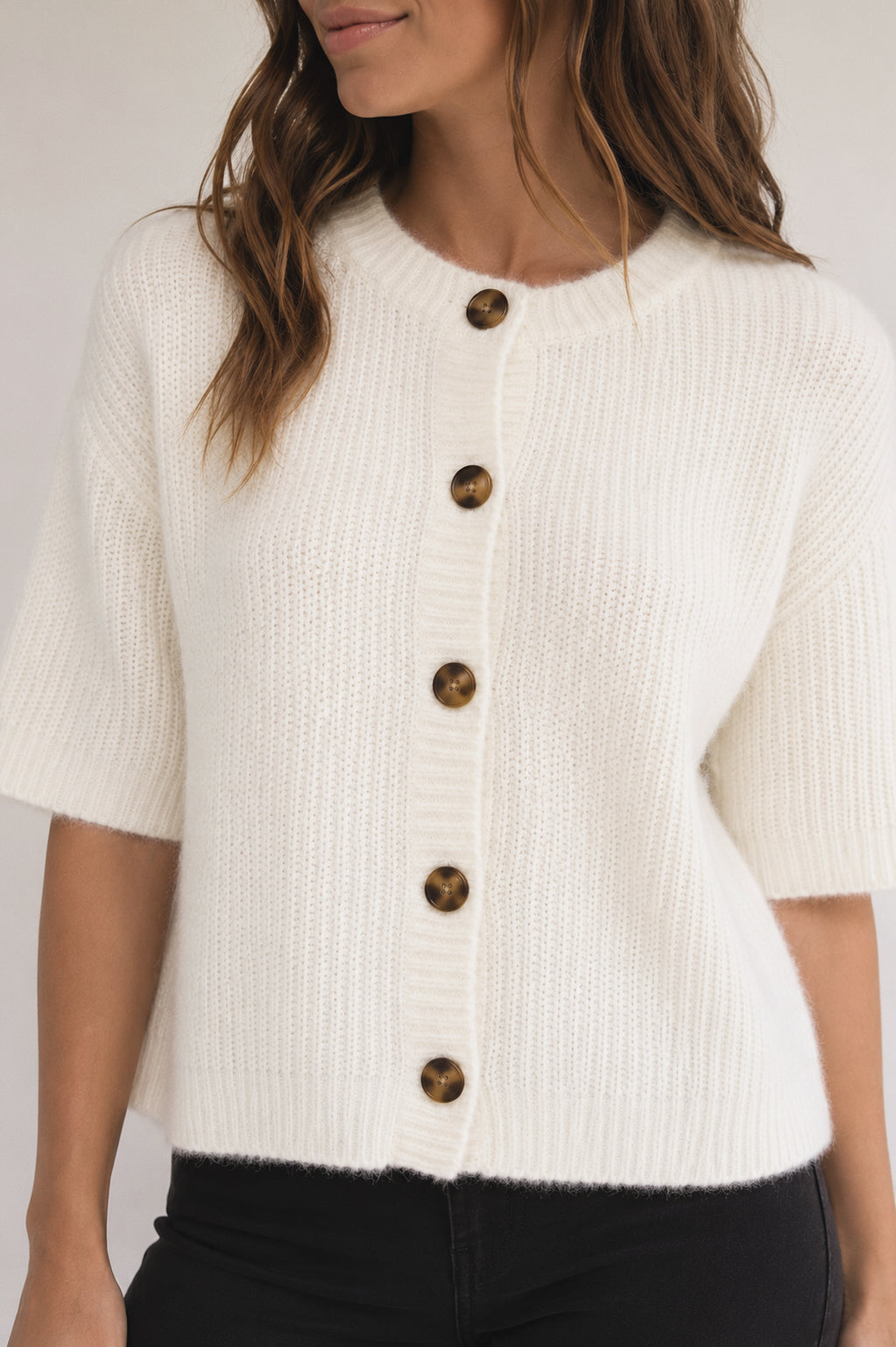 A woman wearing a cream-colored short-sleeve knit cardigan with brown buttons, paired with black pants, against a light gray studio background.