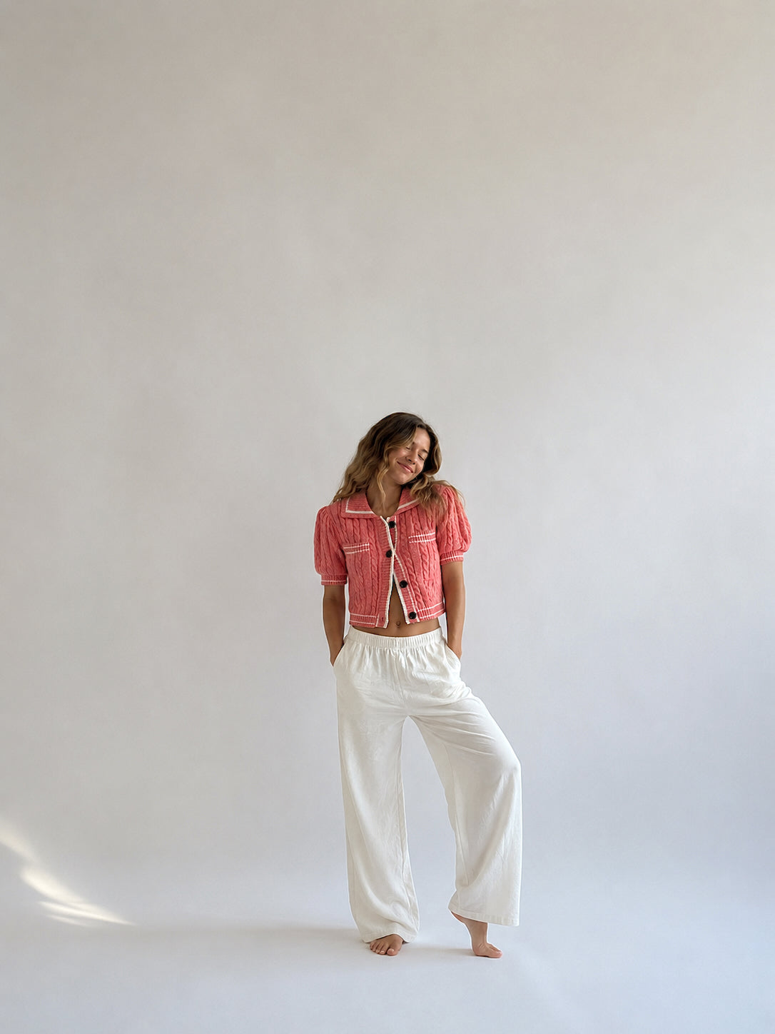A woman posing in a studio setting, wearing a coral pink cropped denim jacket and white wide-leg trousers.