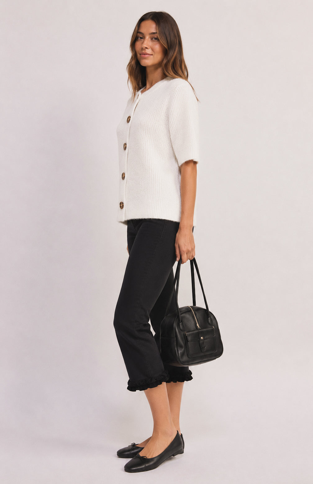 A woman modeling a white short-sleeve button-front cardigan with brown buttons, paired with black cropped pants, black flats, and carrying a black handbag against a light gray studio background.