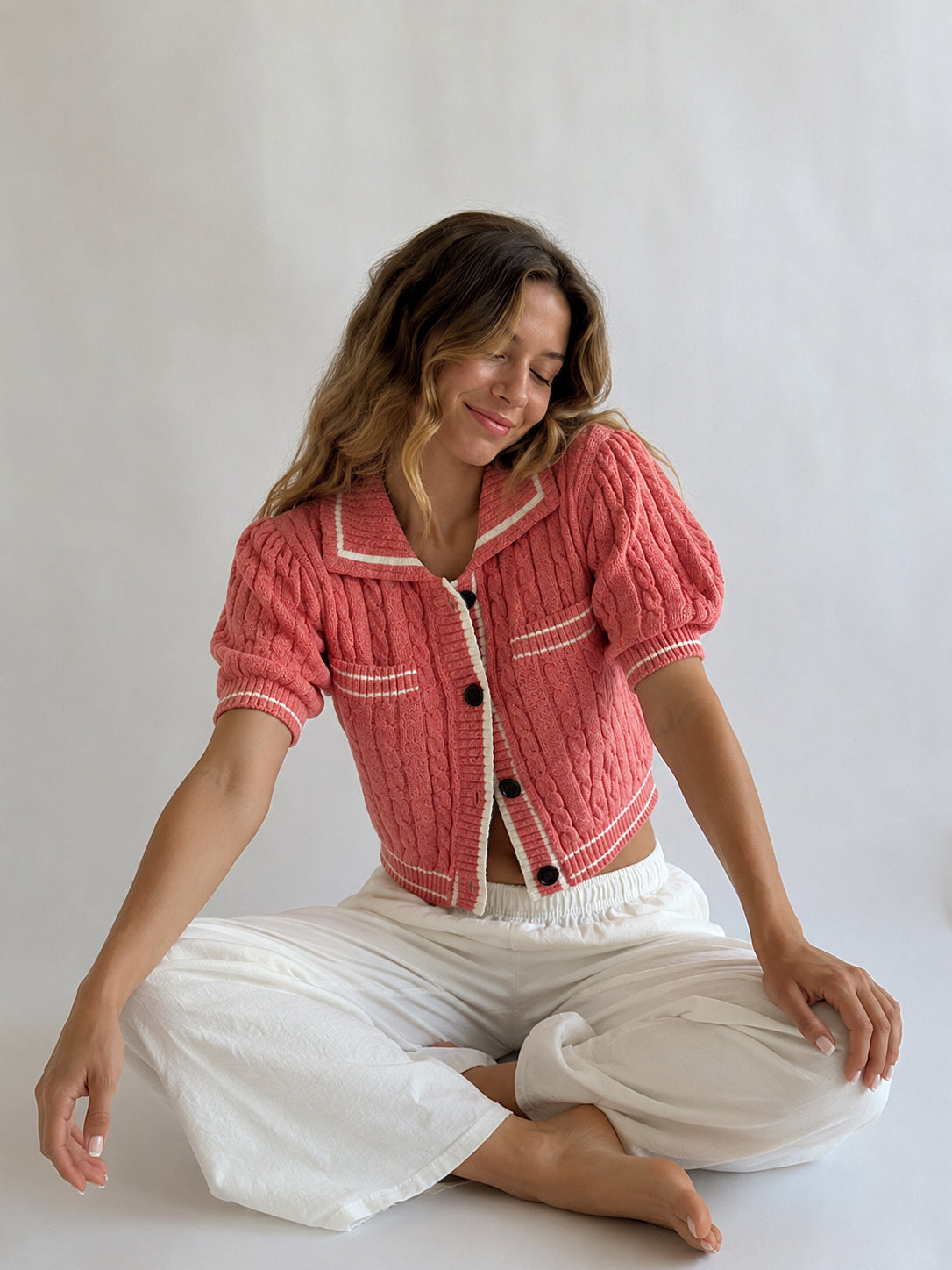A woman sitting cross-legged on a light gray studio floor, wearing a coral pink cable-knit cropped cardigan with white trim and white wide-leg pants.