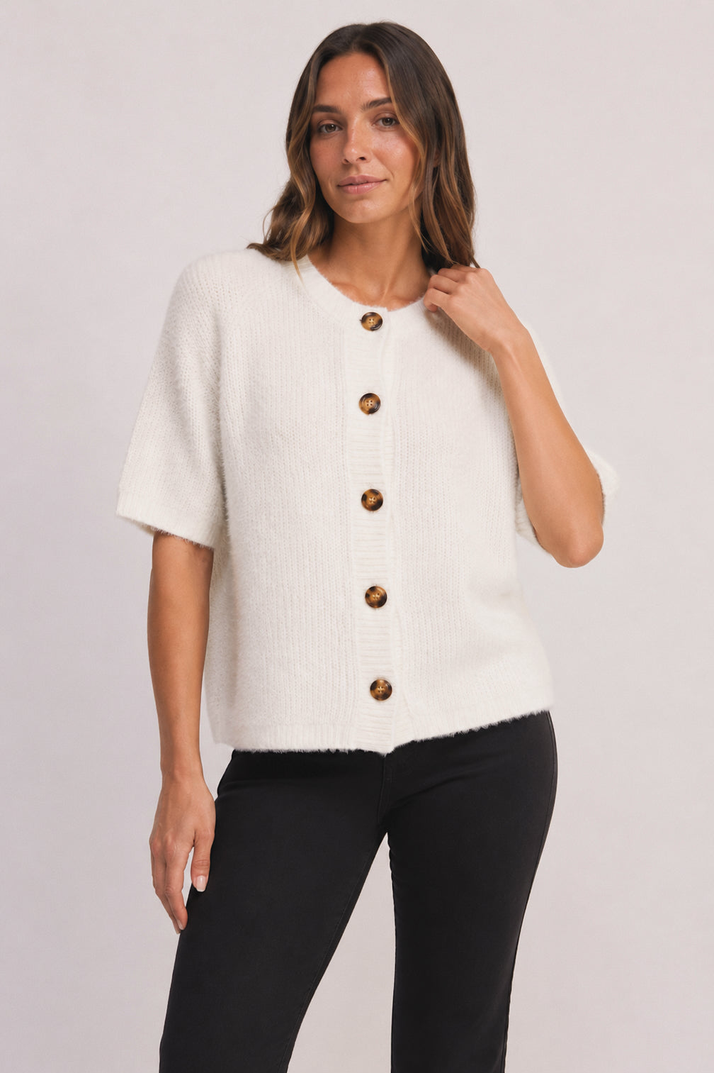 A woman modeling a short-sleeve white knit top with brown buttons, paired with black pants, against a light gray studio background.