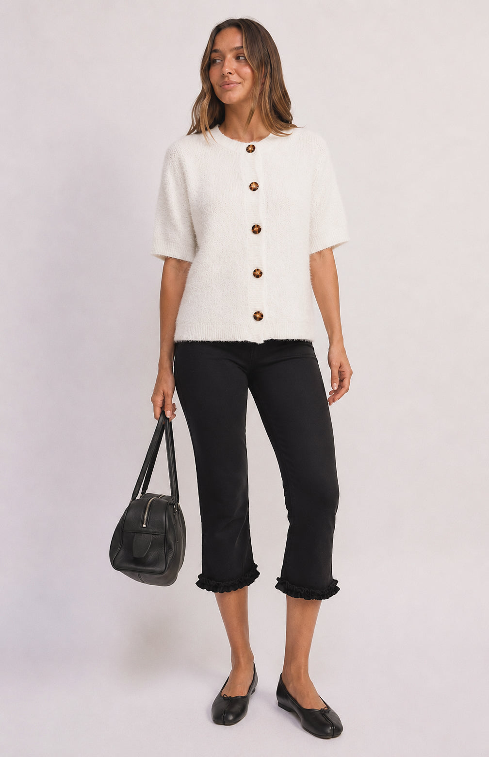 A woman modeling a short-sleeve white textured knit top with brown buttons, paired with black cropped pants and holding a black handbag against a light gray studio background.