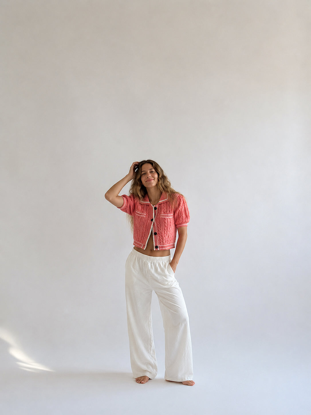 A woman stands barefoot against a plain light gray studio background, wearing a coral-pink cropped knit jacket with white buttons and white wide-leg pants.