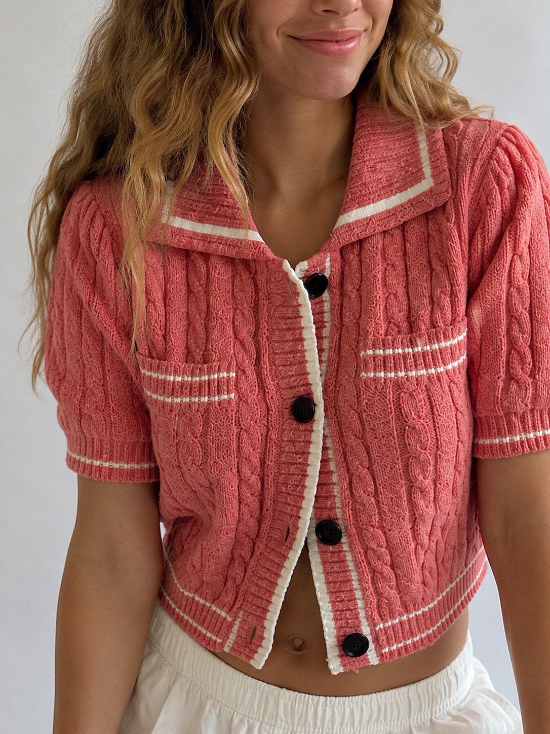 A woman wearing a coral pink cable-knit short-sleeve cropped cardigan with white contrast trim and black buttons, paired with a white skirt.