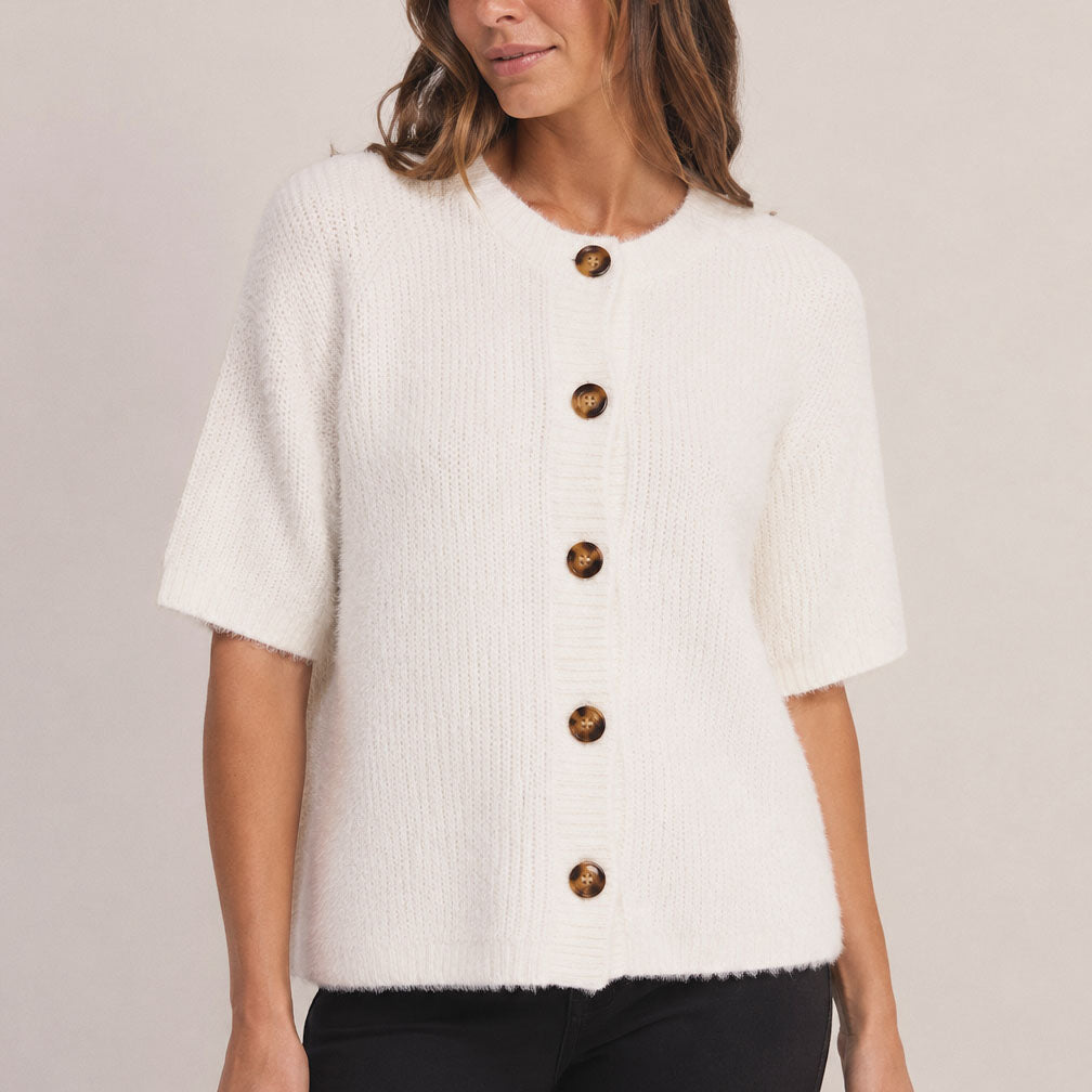 A woman wearing a short-sleeve white textured knit cardigan with large brown buttons, paired with black pants, against a light beige studio background.