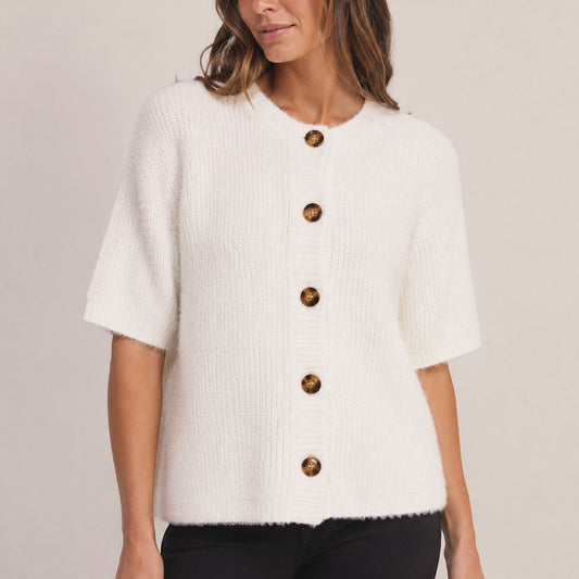 A woman wearing a short-sleeve white textured knit cardigan with large brown buttons, paired with black pants, against a light beige studio background.