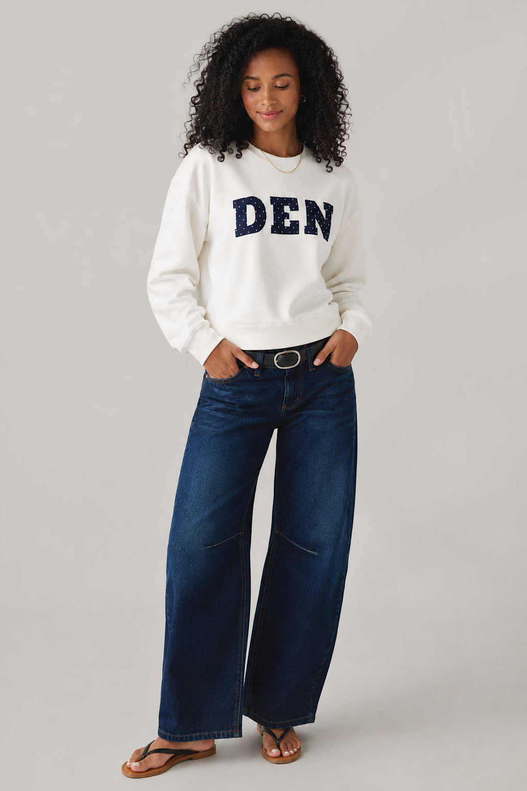 Woman wearing a white sweatshirt with 'DEN' and blue jeans on a gray background