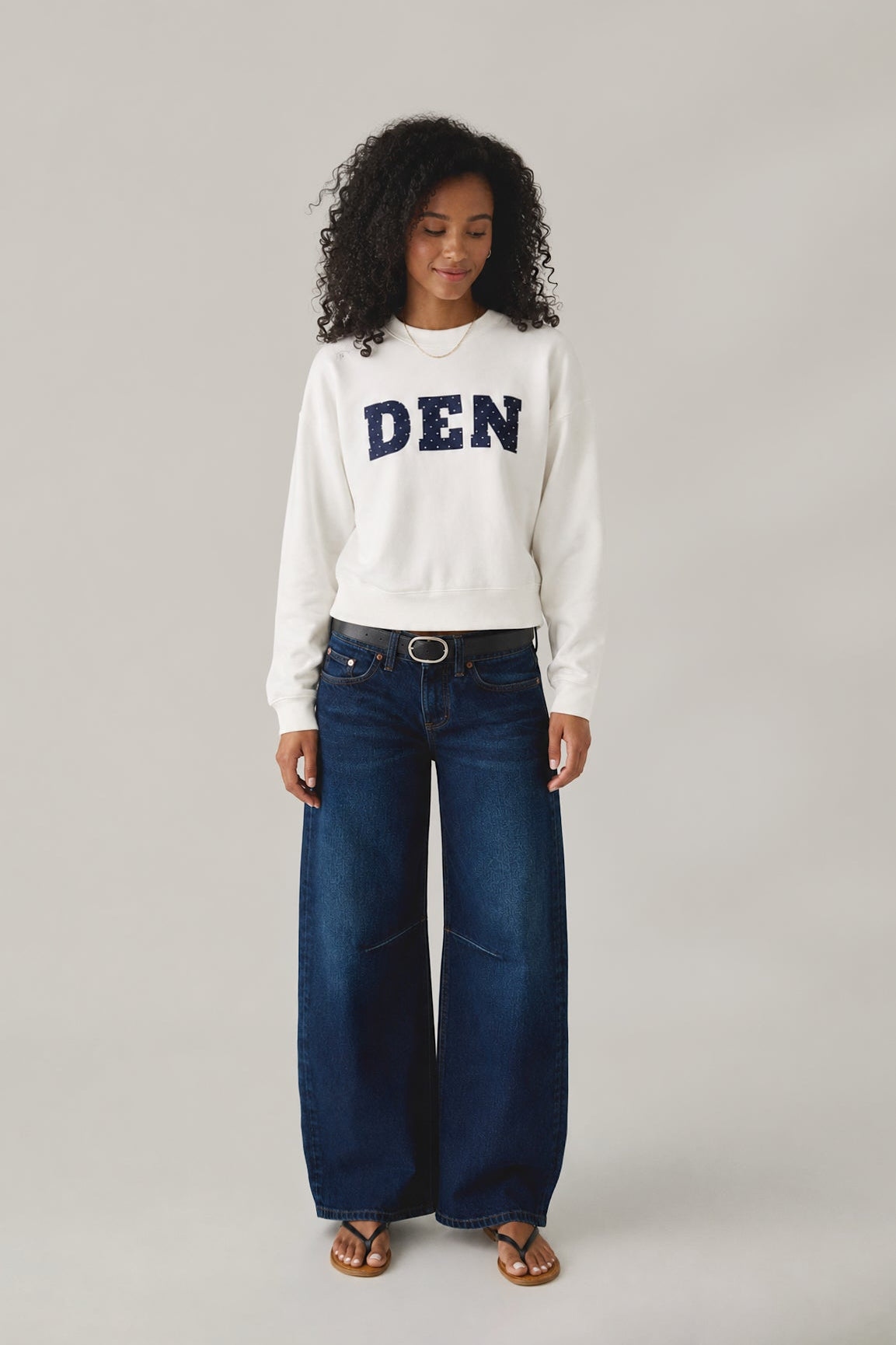 Woman wearing a white sweatshirt with 'DEN' text and blue jeans on a gray background