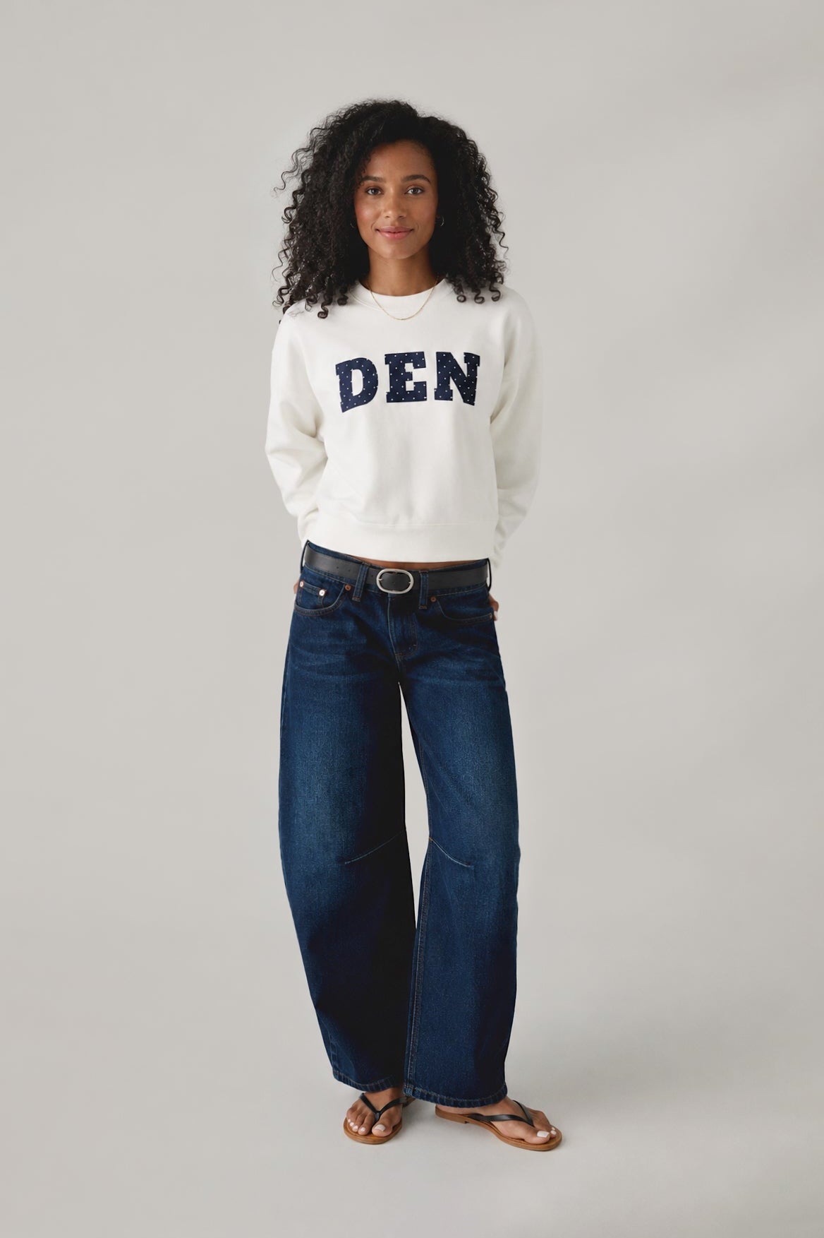 Woman wearing a white sweatshirt with 'DEN' text and dark blue jeans on a gray background
