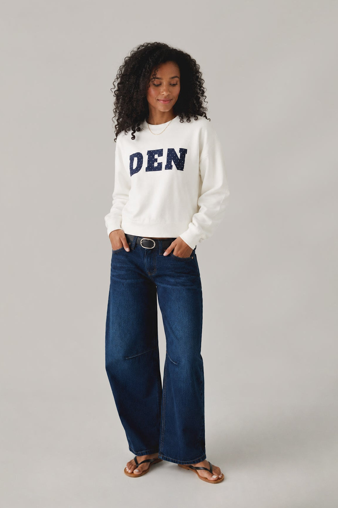 Woman wearing a white sweatshirt with 'DEN' text and blue jeans on a gray background