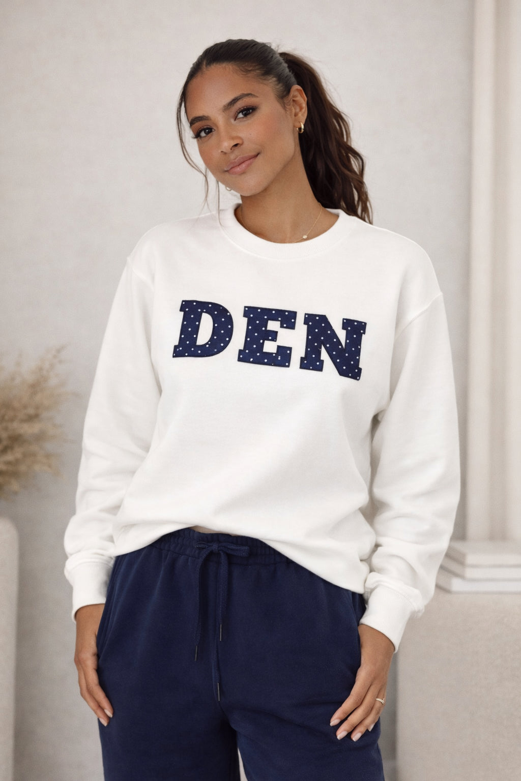 Woman wearing a white sweatshirt with 'DEN' in blue letters and navy pants.