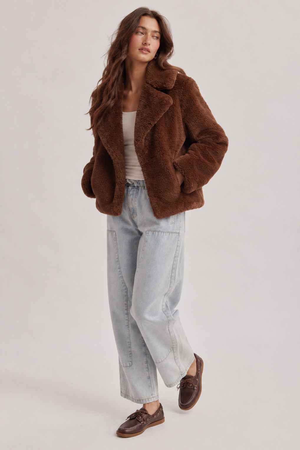 A woman modeling a brown teddy fleece jacket, styled with light-wash wide-leg jeans, a white tank top, and brown leather loafers, against a light gray studio background.