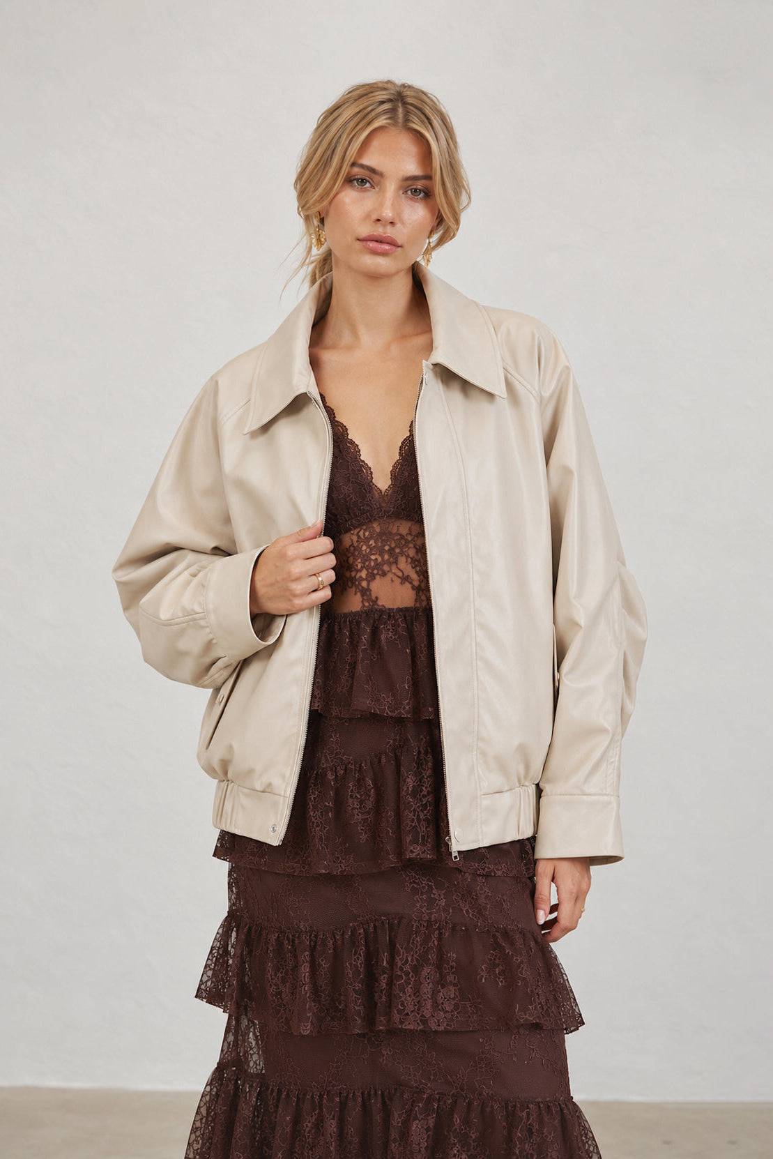 A woman modeling a light beige leather jacket over a dark brown lace dress, standing against a plain light gray wall.