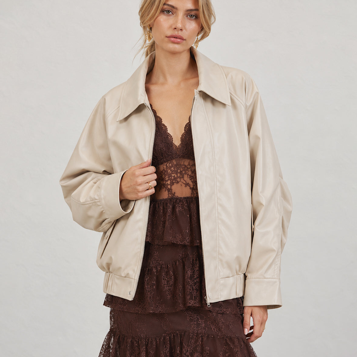 A woman wearing a light beige faux-leather jacket over a dark brown lace dress, standing against a light gray textured wall.