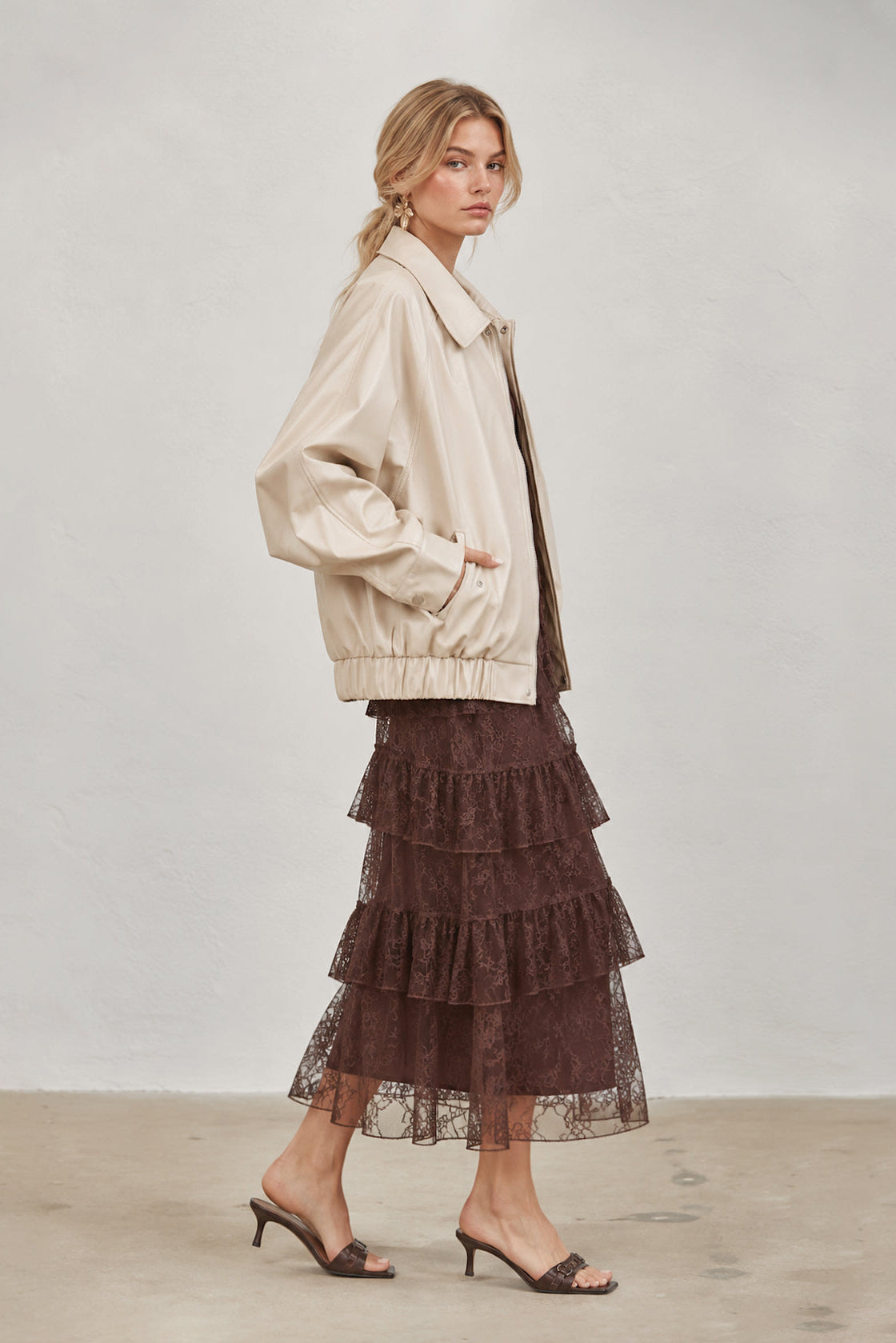 A woman models a cream-colored cropped jacket paired with a dark brown tiered lace maxi skirt and brown heeled sandals, standing against a neutral light gray wall.