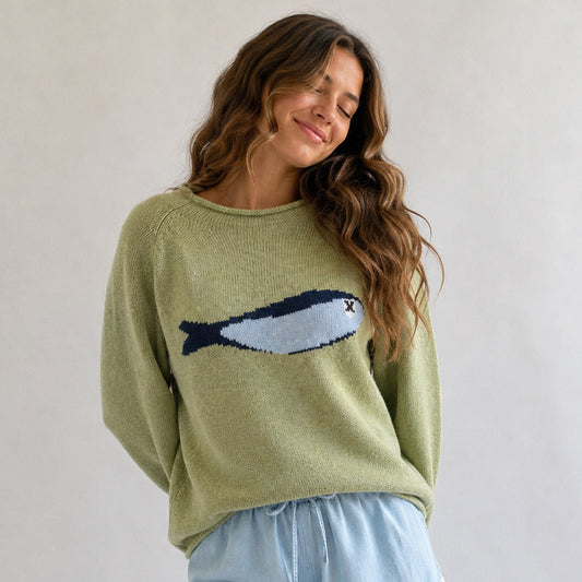 A woman wearing a light olive green knit sweater with a knitted fish motif across the chest, paired with light blue pants, set against a plain light gray background.